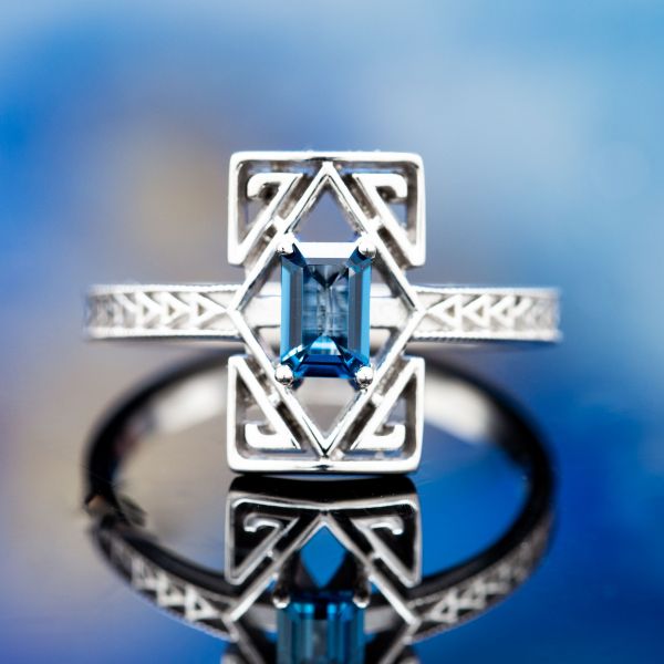 A stunning geometric design with Art Deco inspiration, surrounding an emerald cut London blue topaz with diamonds and spiraling triangles and a repeating triangle design on the shank.