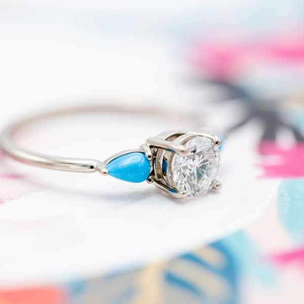 A delicate three-stone engagement ring surrounds a perfect diamond center stone with bright pops of turquoise curving into the white gold band.