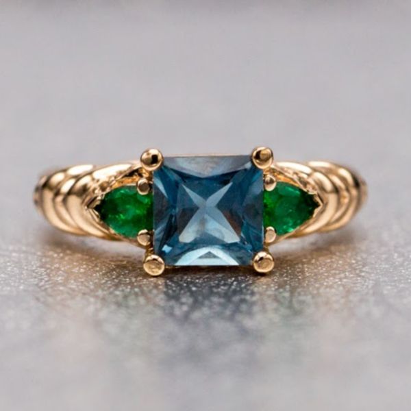 This Art Nouveau-inspired design sets a square cut London blue topaz between pear cut emeralds and highlights the side profile's organic rose gold curves with Swiss blue topaz accents.
