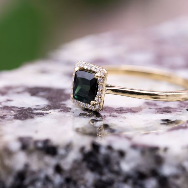 A rich, green cushion cut green tourmaline sits in a floating diamond halo in this classic gold ring.